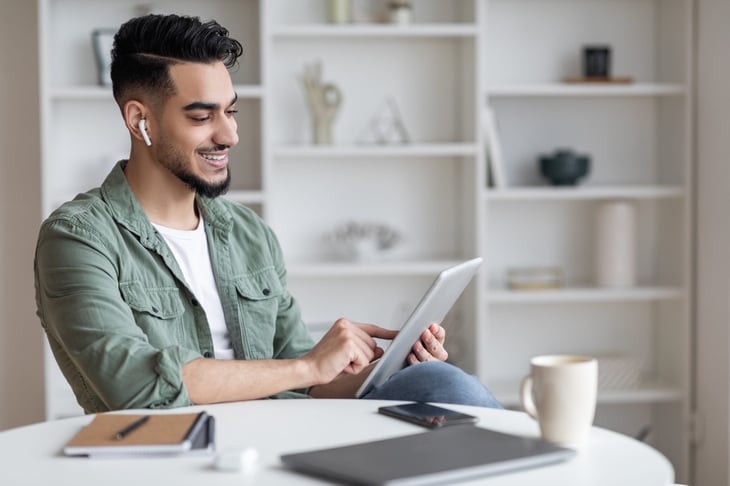 Man using Airpods and tablet at home with a coffee mug and laptop on the table.